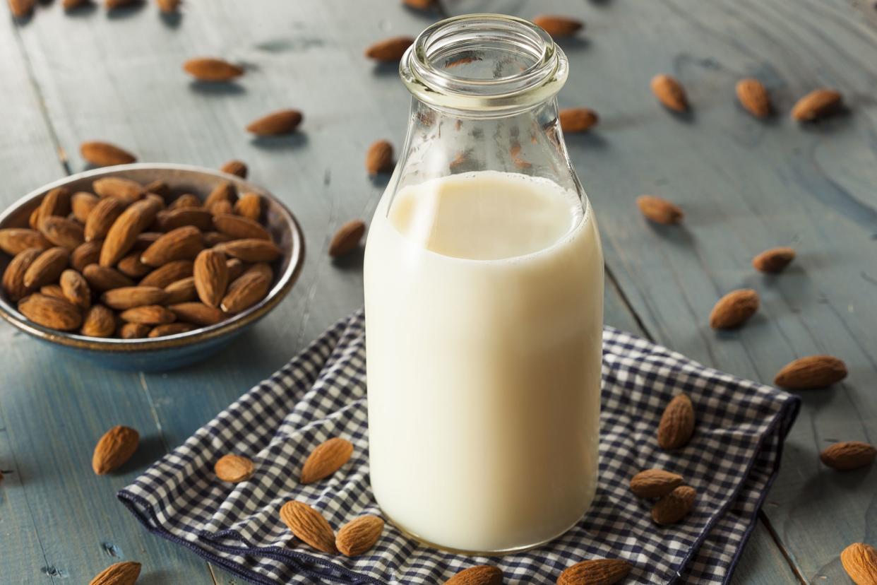 Almond milk in an opened glass jug on a blue checkered napkin surrounded by almonds on a blue wooden table