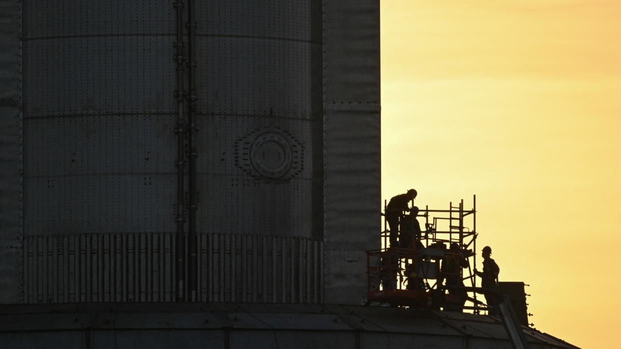  Workers on scaffolding in hard hats next to a giant metal rocket. 