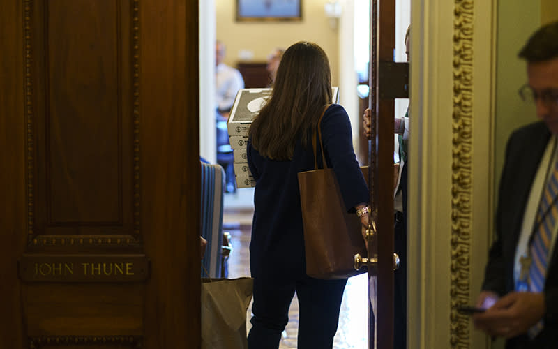 An aide is shown from behind walking through a doorway and carrying a stack of five pizza boxes