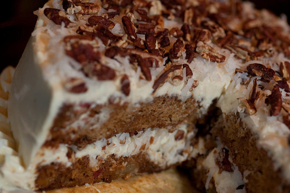 A close-up view of a slice taken out of a layered carrot cake topped with shredded coconut and chopped nuts