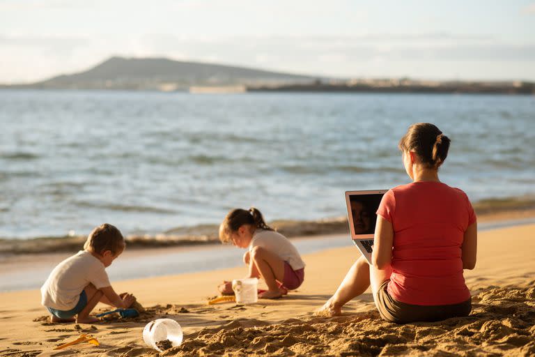 Rear shot of mother working remotely as kids play on the beach