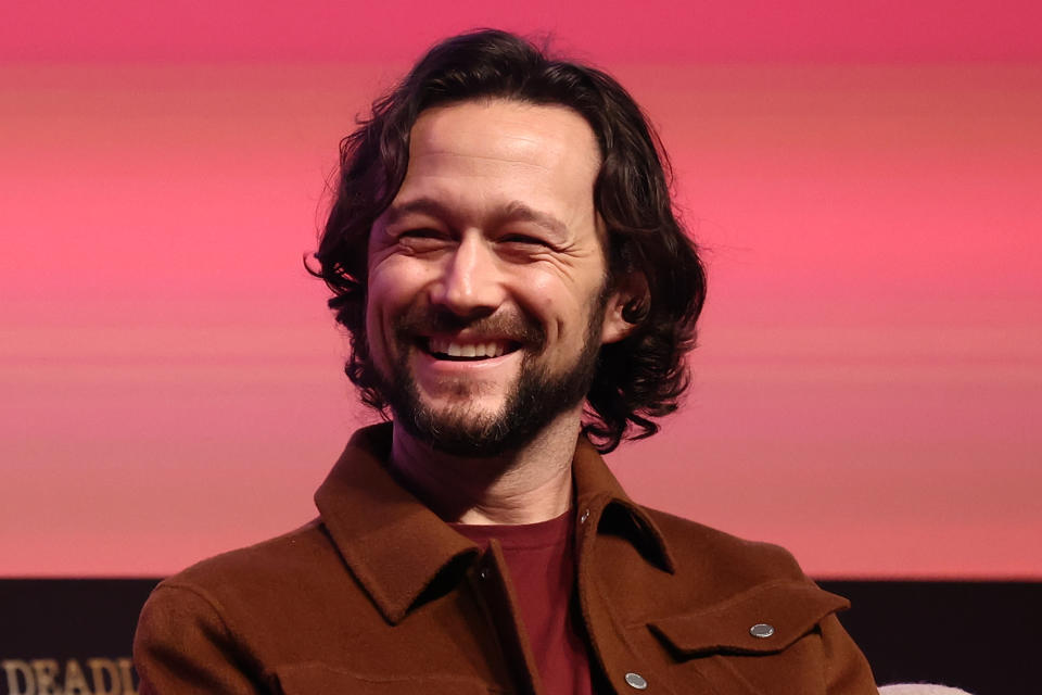 Joseph Gordon-Levitt is smiling sat in front of a pink background.