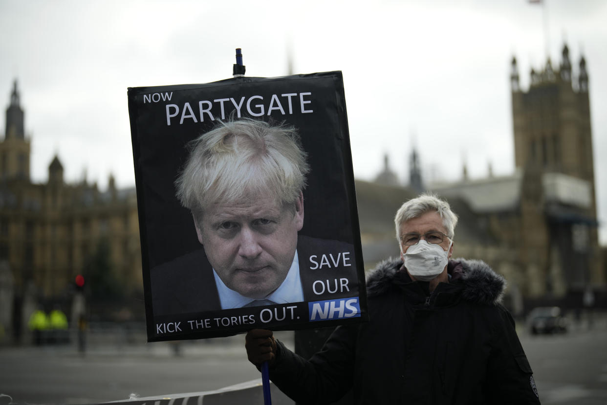 An anti-Conservative Party protester holds a placard with an image of British Prime Minister Boris Johnson including the words "Now Partygate" backdropped by the Houses of Parliament, in London, Wednesday, Dec. 8, 2021. British Prime Minister Boris Johnson on Wednesday ordered an inquiry and said he was "furious" after a leaked video showed senior members of his staff joking about holding a lockdown-breaching Christmas party. (AP Photo/Matt Dunham)
