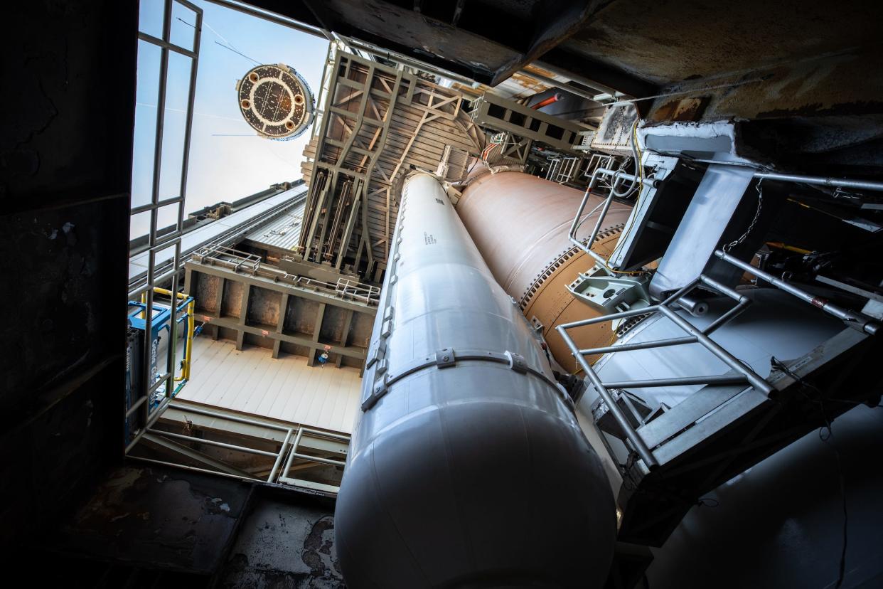 view from the bottom of a rocket looking up, inside an open-air facility with wooden scaffolding around the rocket and the round bottom of a spaceship visible in the corner being lifted toward the top of the rocket