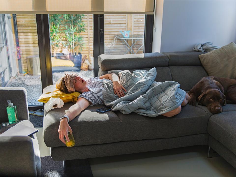 A woman is lying on her couch under a blanket with a brown washcloth over her forehead. She is kept company by her chocolate Labrador, who is curled up at her feet. Her outstretched hand holds a nearly empty glass of orange juice.