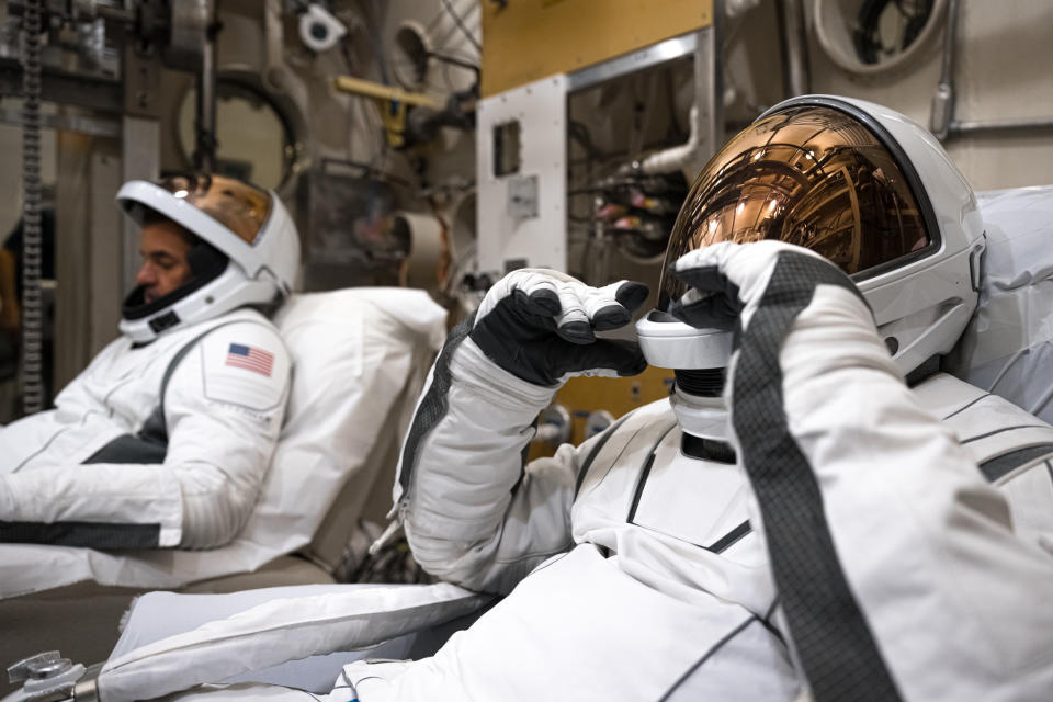 Two astronauts, one in the foreground and one in the background, both facing left, are reclining in white and black bucket suits and white space suits.