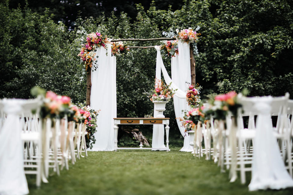 A beautifully decorated outdoor wedding altar with floral arrangements and white drapery, set between rows of chairs on a grassy lawn