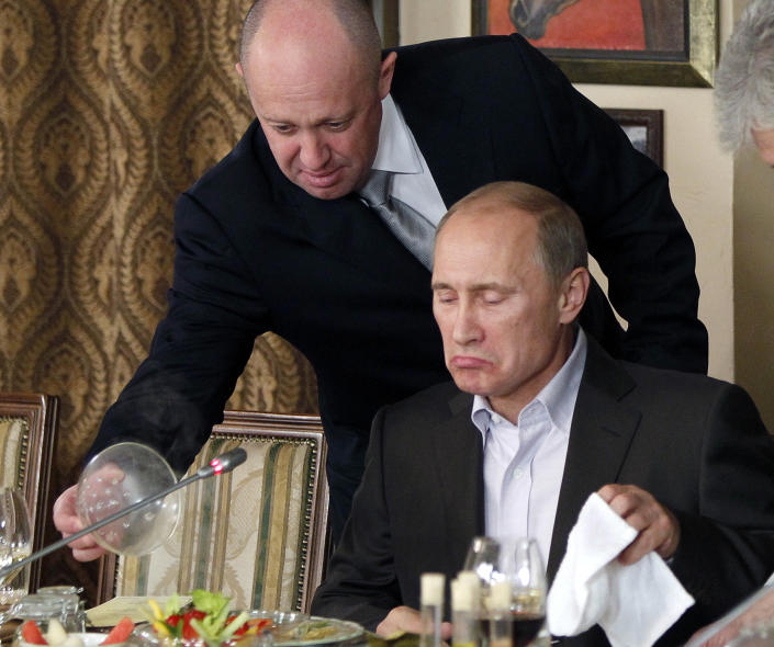 Yevgeny Prigozhin, standing, lifts a lid from a plate of food in front of seated Vladimir Putin as Putin makes a judgmental face as though he's trying to determine whether it will be to his liking.