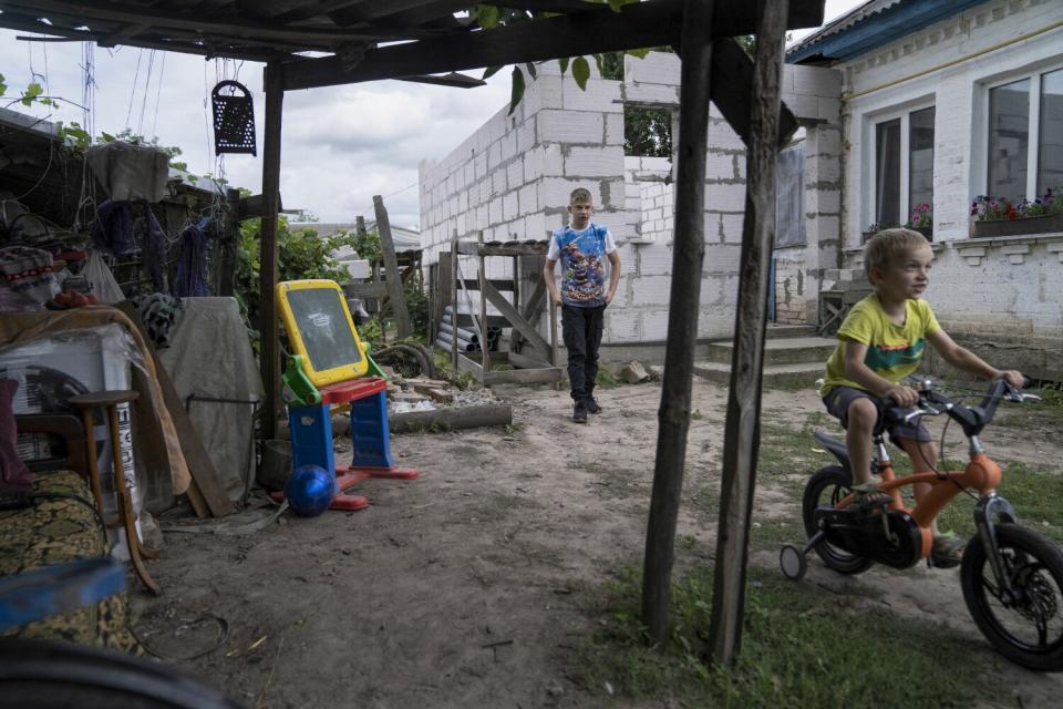 A boy rides a bicycle as an older boy watches in a yard.