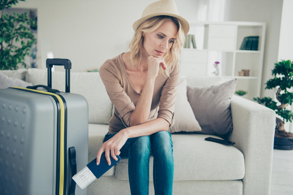 Gorgeous attractive lady in her casual clothes denim jeans blond curly wavy hair she sit on cozy comfort couch sofa inside modern apartment room hold boarding pass in hands look sad