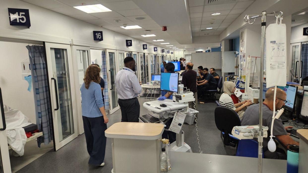 A busy hospital ward filled with staff and patients, there are patient bays running down the left side of the ward and to the right staff are seen working on computers