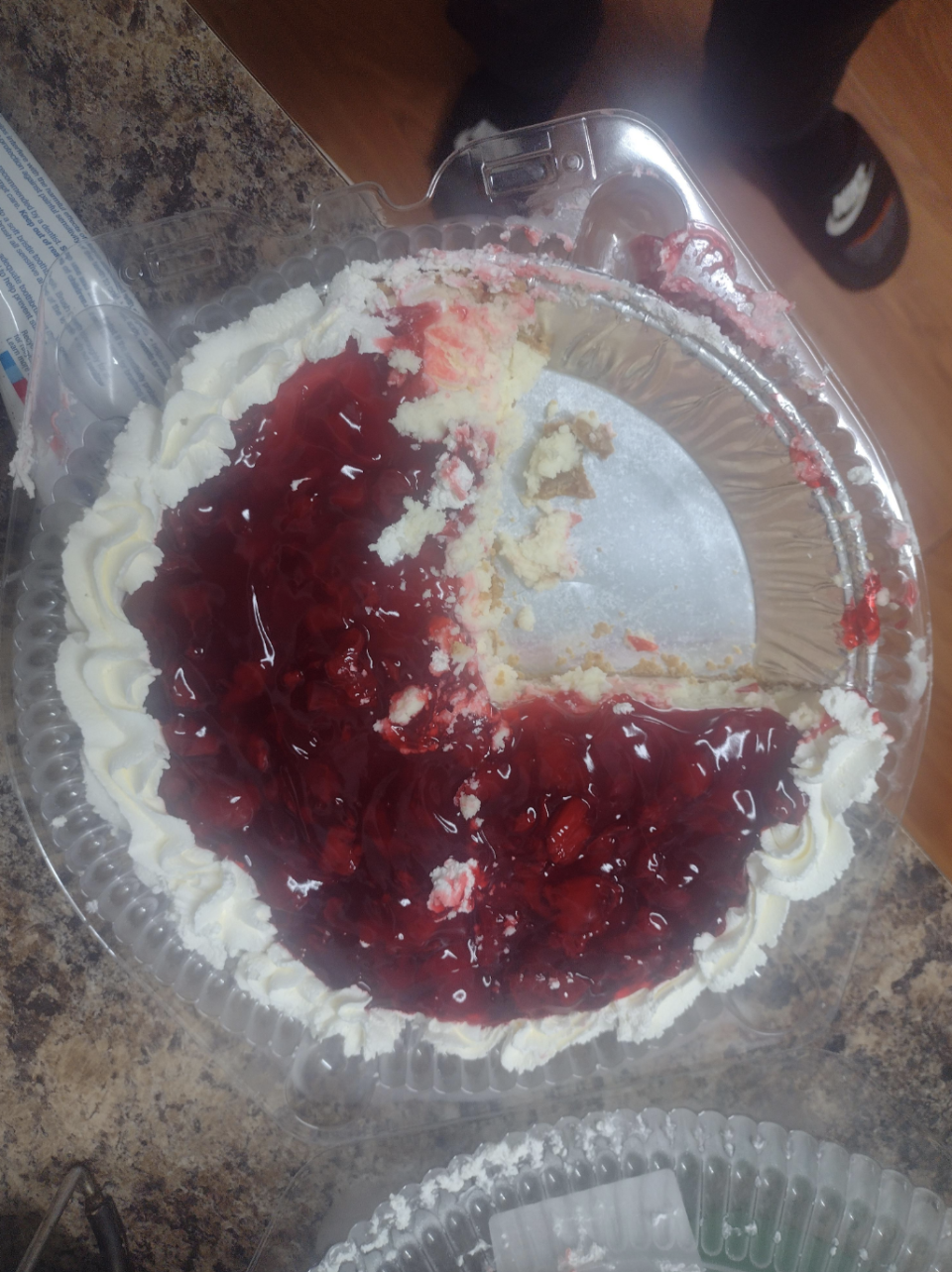 A partially eaten cherry-topped cheesecake in a plastic container sits on a countertop. Someone's feet are visible in the background