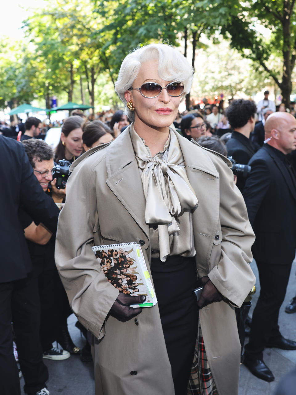 Alexis Stone, styled as Miranda Priestly, wearing a trench coat, silk blouse with a bow, and sunglasses, holding a notebook, amidst a crowd outdoors