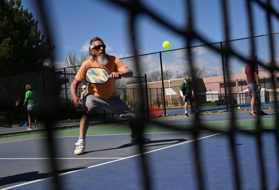 A bearded pickleball player wears sunglasses as he prepares to hit a bright yellow pickleball on a sunny day.  The entire scene is seen through a net.