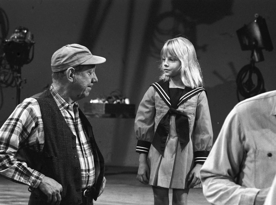 paul hartman and jodie foster stand and look at each other with video equipment in the background, he wears a plaid shirt, vest and hat, she wears a nautical dress