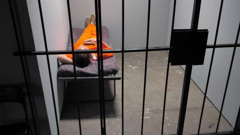 A single prisoner in an orange jumpsuit lies on a cot in a prison cell.