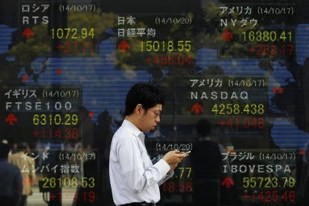 A pedestrian uses his mobile phone as he walks past an electronic board showing the stock market indices of various countries outside a brokerage in Tokyo October 20, 2014. REUTERS/Yuya Shino