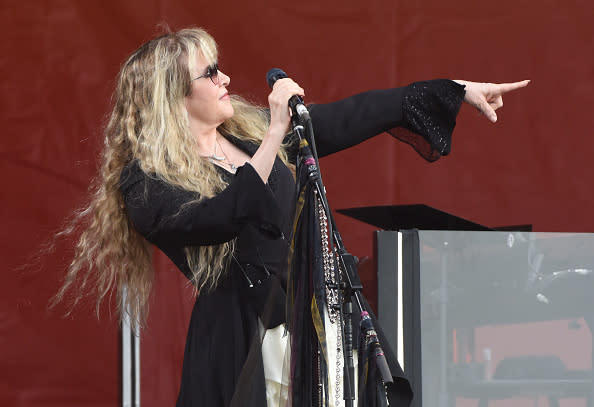 Stevie Nicks performs on stage, wearing flowing bohemian-style clothing and pointing towards the audience