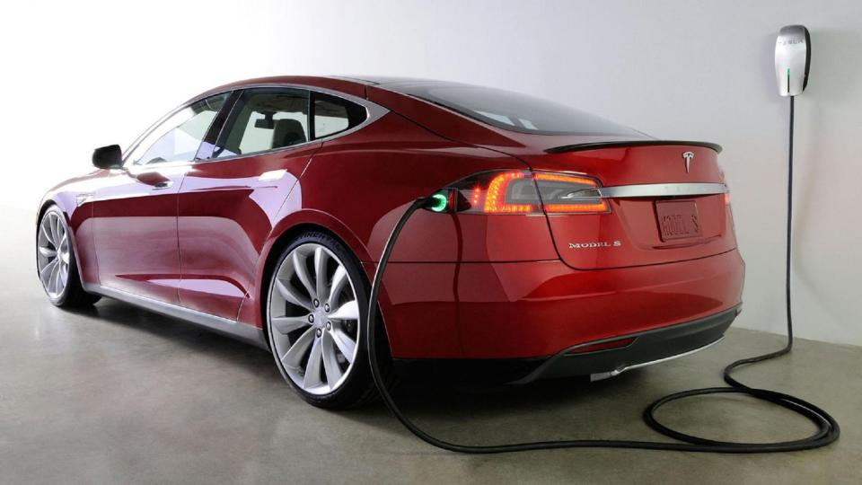 An all-electric Tesla Model S sedan plugged into a wall outlet for charging.