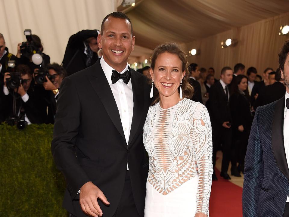Alex Rodriguez stands in a suit and Anne Wojcicki stands in a white dress on a red carpet.