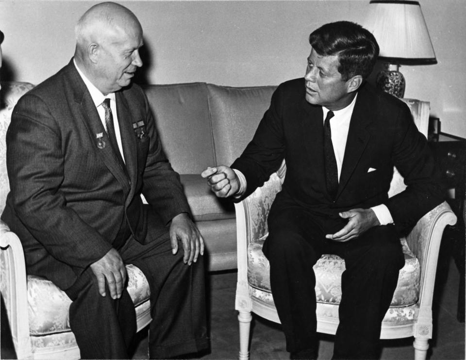 Black and white photo of two men sitting in chairs wearing dark suits and ties.  The man on the left is bald.