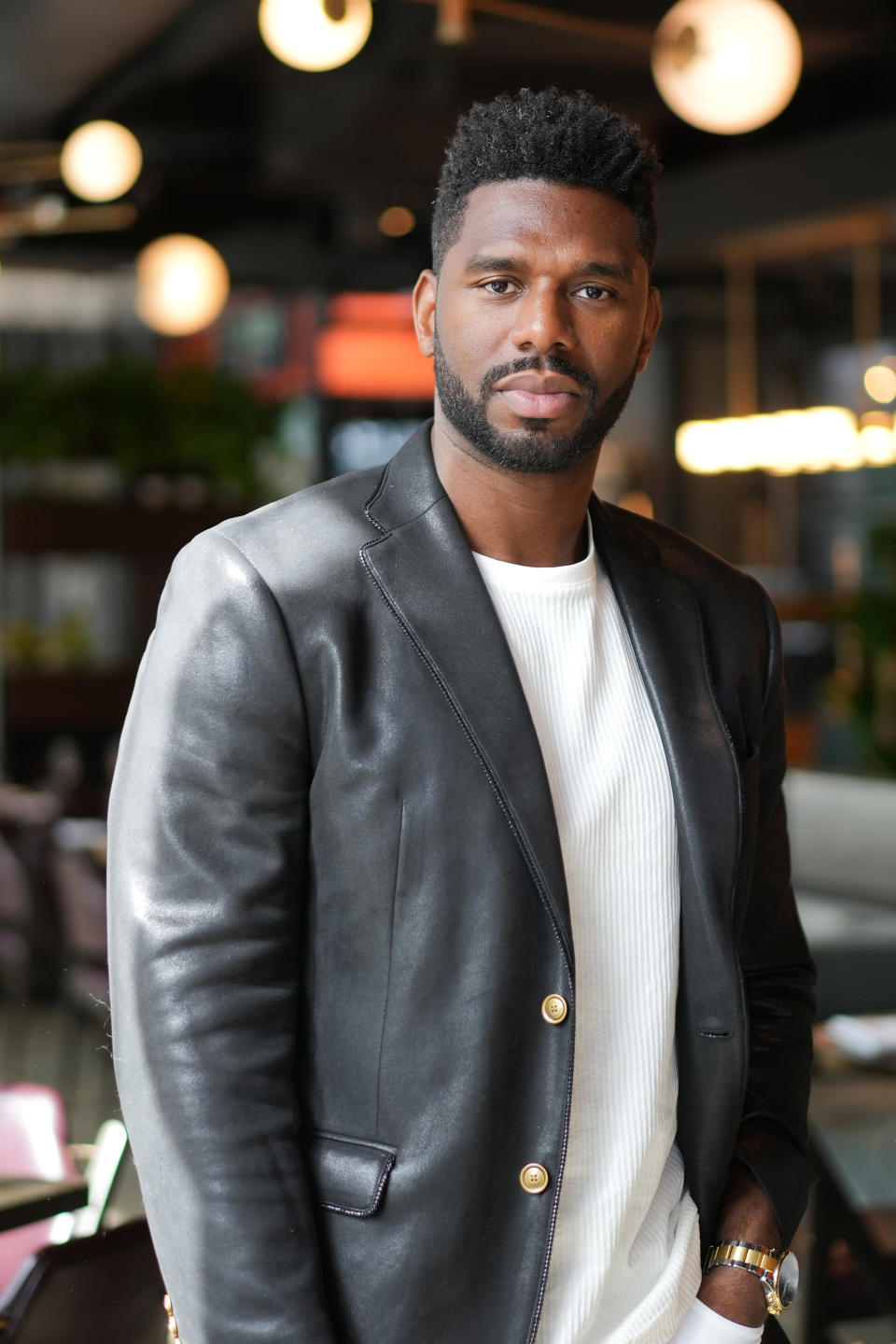 Man in leather jacket and white shirt, standing indoors, looking at camera