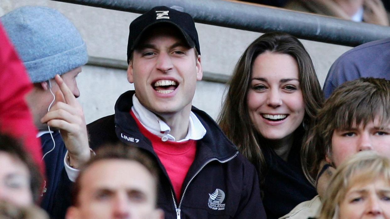 Prince William in a black hat and jacket with a red shirt laughs at a match with Kate Middleton in black