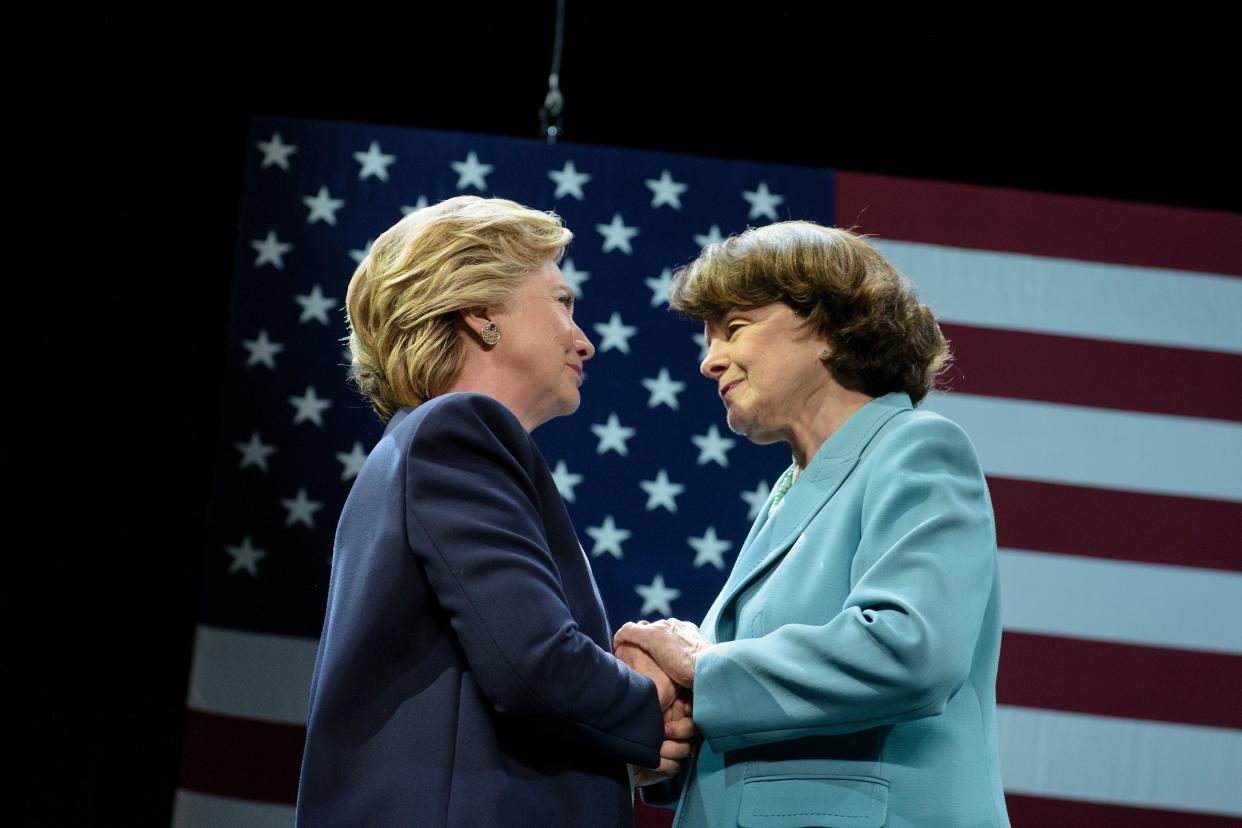 Hillary Clinton and Dianne Feinstein clasp each others' hands in front of an American flag.
