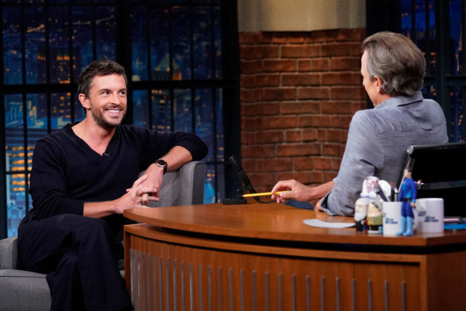 Jonathan Bailey and Seth Meyers on a talk show set, sitting and conversing. McNally is dressed casually, while Meyers wears a suit