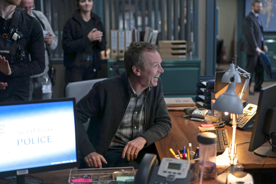 Steven Robertson laughing in a behind-the-scenes picture taken on the Shetland police station set, with crew members and cameras visible in the background