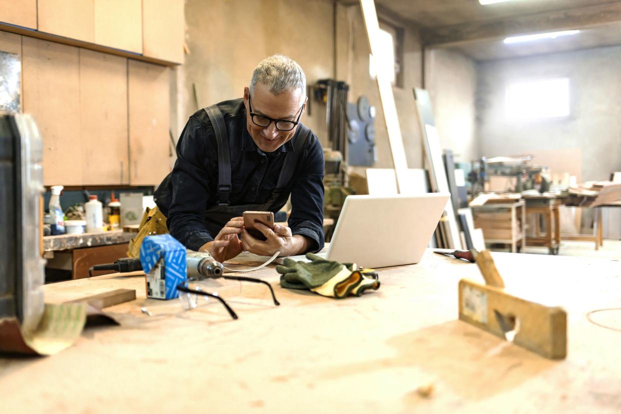 senior man using laptop and texting on smartphone in his carpentry workshop