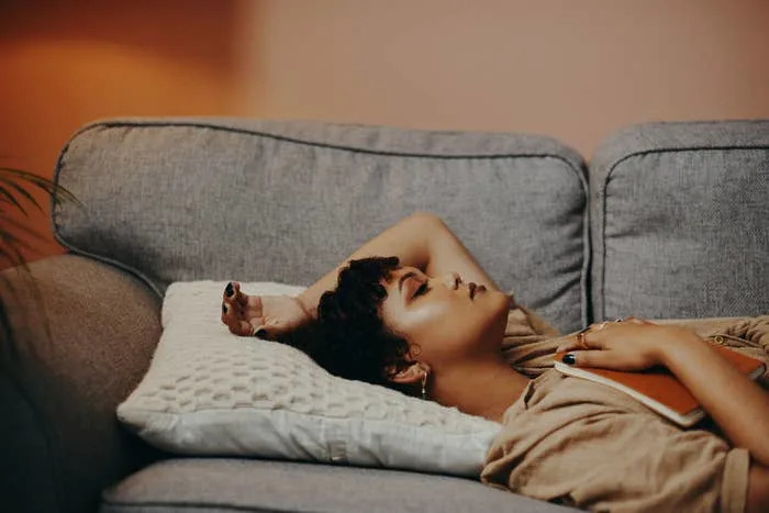 A woman lying on her couch with her eyes closed, one arm raised behind her head, and a book resting on her chest