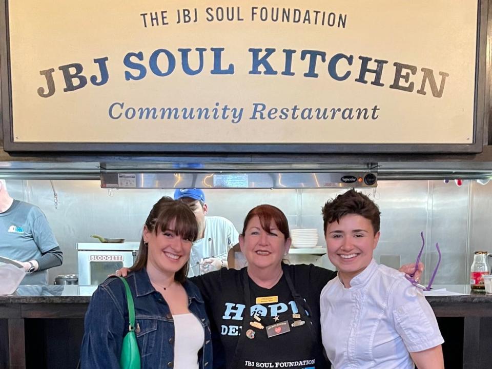 A large sign with the words "The JBJ Soul Foundation JBJ Soul Kitchen Community Restaurant." It's surrounded by dark wood shelving with colorful merchandise and awards. Below the sign, there is an opening into the kitchen showing cooks. Victoria is smiling with a server and a chef in front of it.