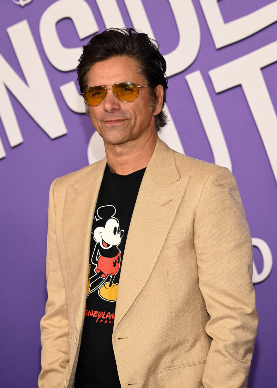 John Stamos wearing a Mickey Mouse T-shirt and beige blazer, attending a celebrity event in front of a large purple "Inside Out" sign
