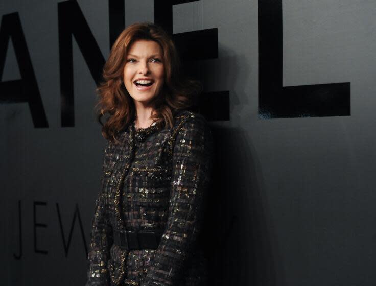 Linda Evangelista smiles while wearing a black patterned coat and skirt against a black backdrop.