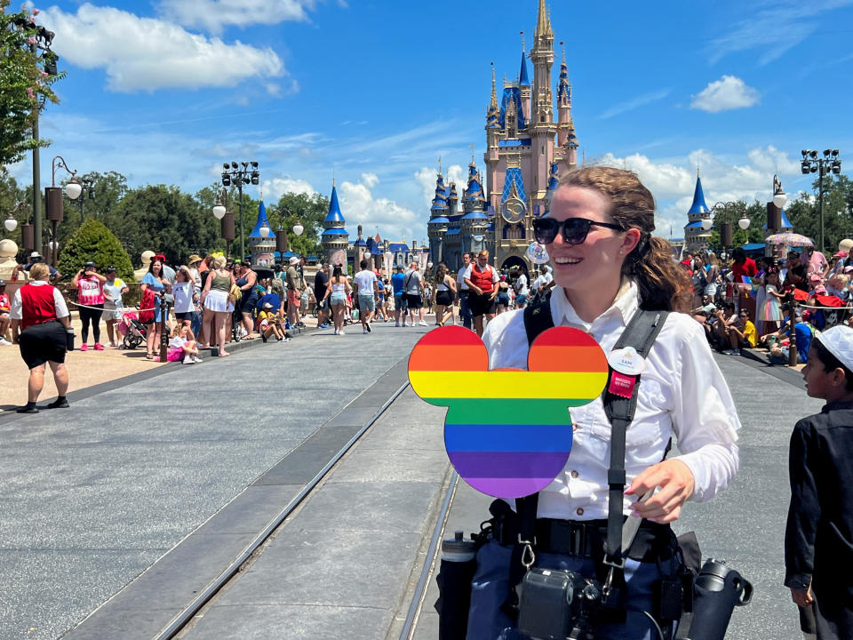 A young woman holds a rainbow-colored Mickey Mouse cutout, with the Magic Kingdom in the background. 