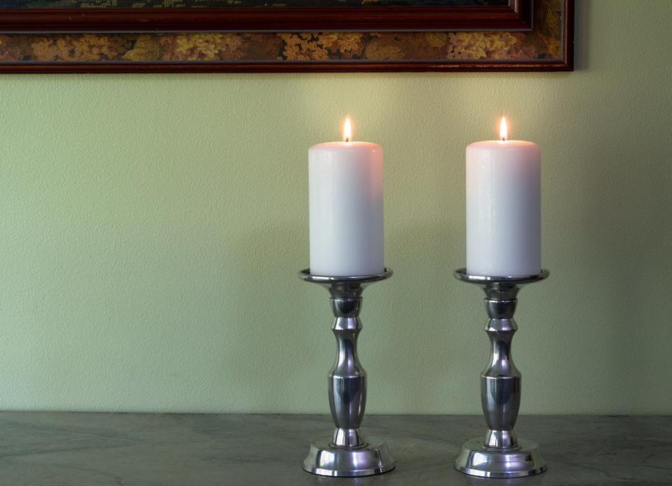 Two lit white candles in candle holders in front of a wall painted light green.