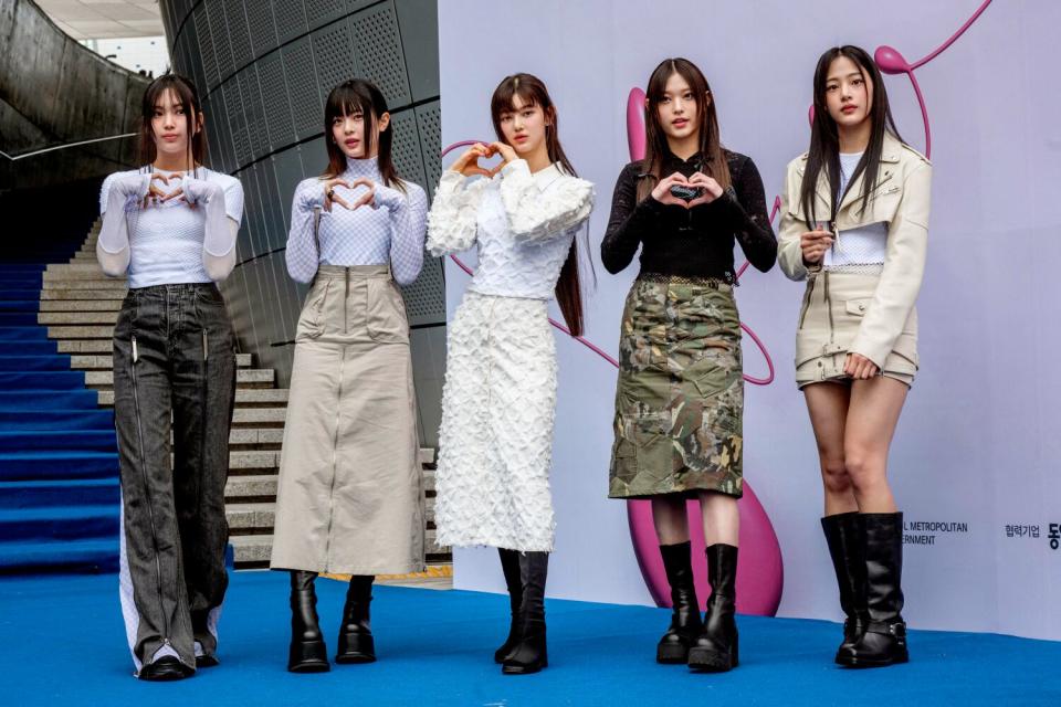 Five members of South Korean K-pop group NewJeans stand making hearts with their hands for a photo