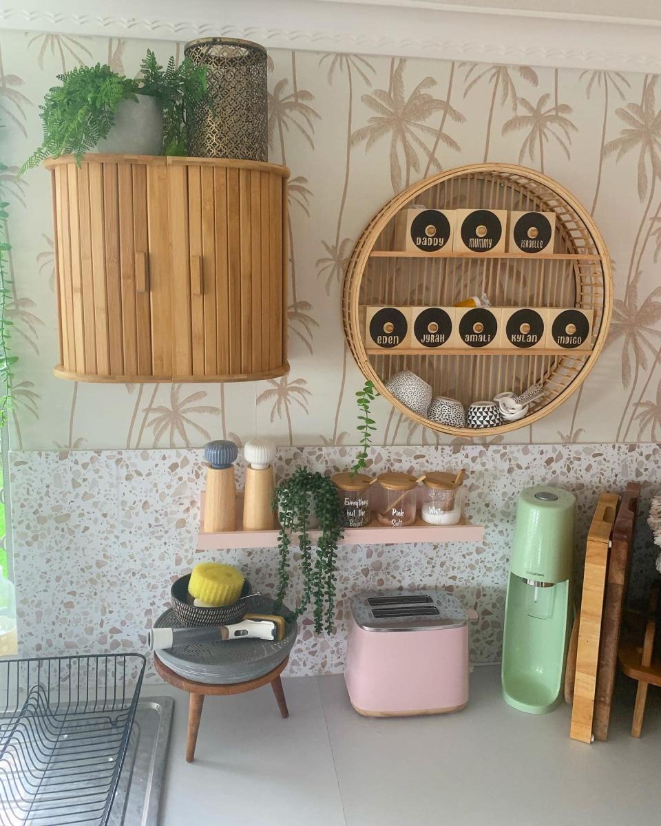 A wall cabinet made out of two Kmart bread bins joined together sits on a palm-patterned wall, next to a round rattan shelf and plants in a kitchen scene including a bench with toaster, drink mixer and sink rack.. 