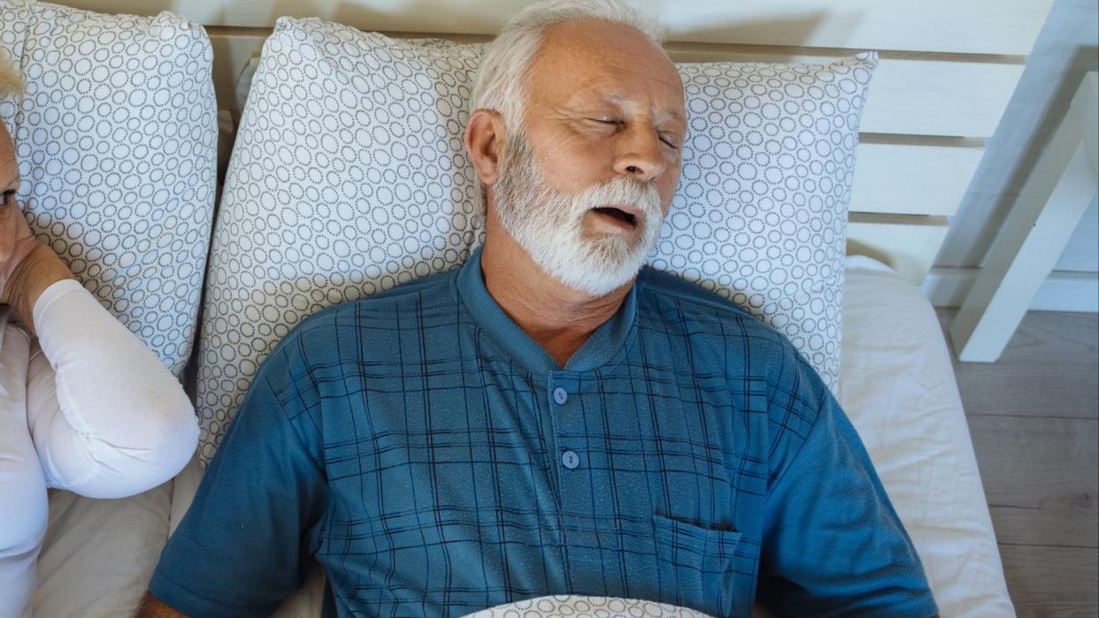  photo of an older man with white hair and a blue shirt sleeping in bed. his mouth is slightly open and a woman can be seen lying near him covering her ears as if he's snoring 