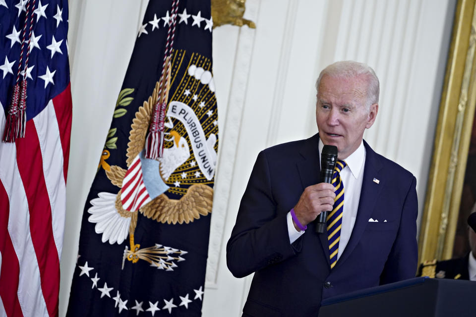 President Biden stands at a podium holding a microphone next to two flags.