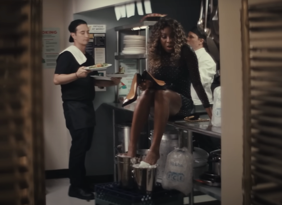A woman in sparkly dress cools her feet in ice buckets in a restaurant kitchen, while a waiter looks on with a tray of food