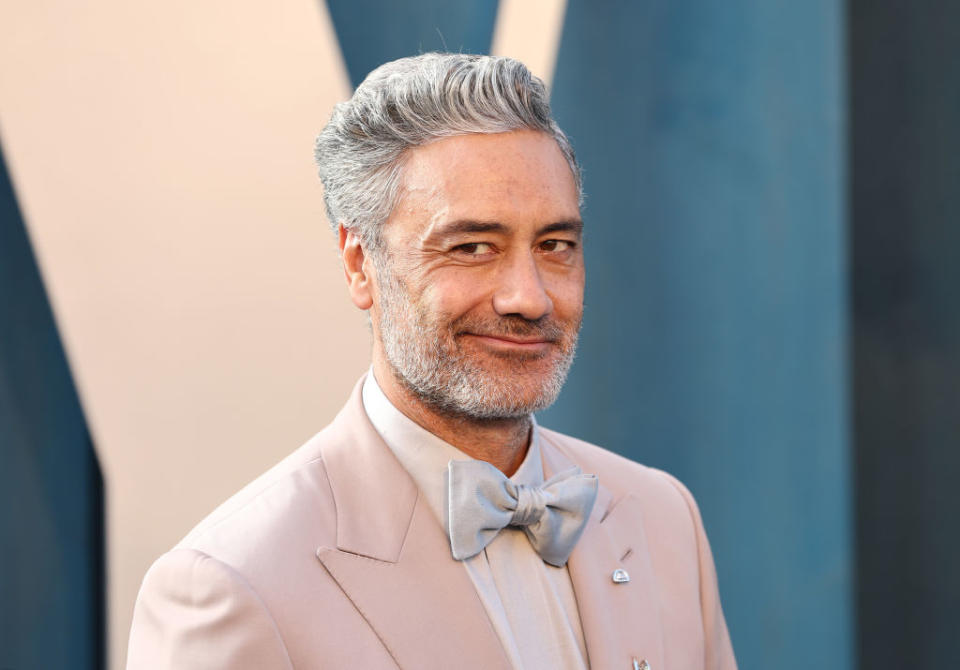 Taika Waititi at an event, wearing a light suit with a bow tie and a small metallic pin on his lapel, smiling at the camera