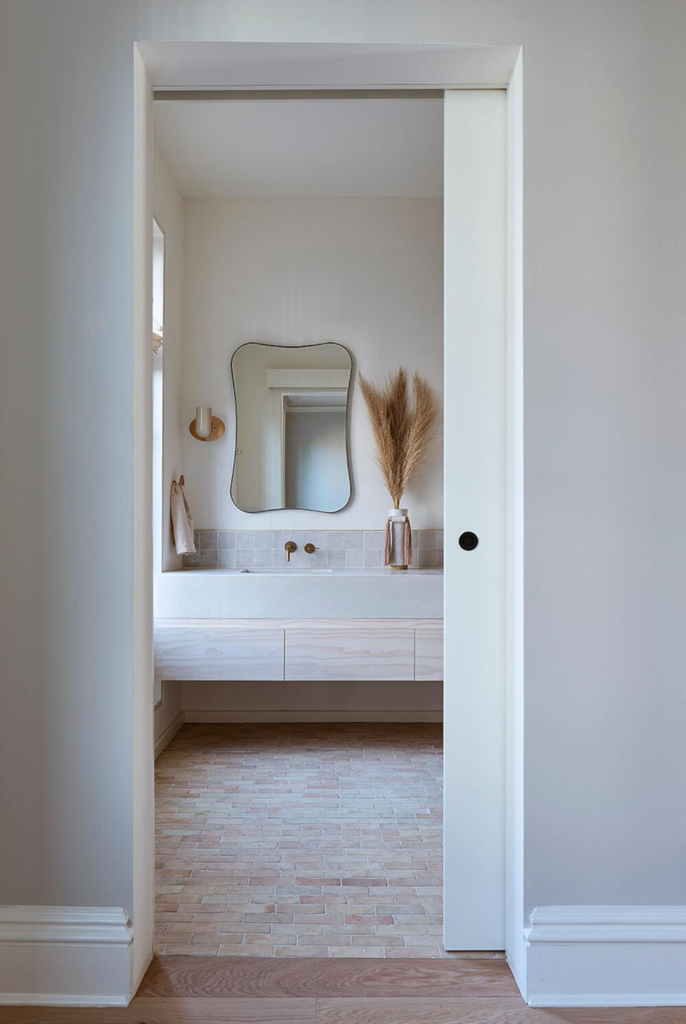 Through a pocket door, view a bathroom with warm white walls, a wall-mounted vanity, and a pale brick floor.