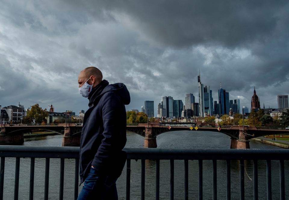 A man wearing a face mask walks over a bridge with the buildings of the banking district in background in Frankfurt, Germany, Thursday, Oct. 29, 2020. (AP Photo/Michael Probst)