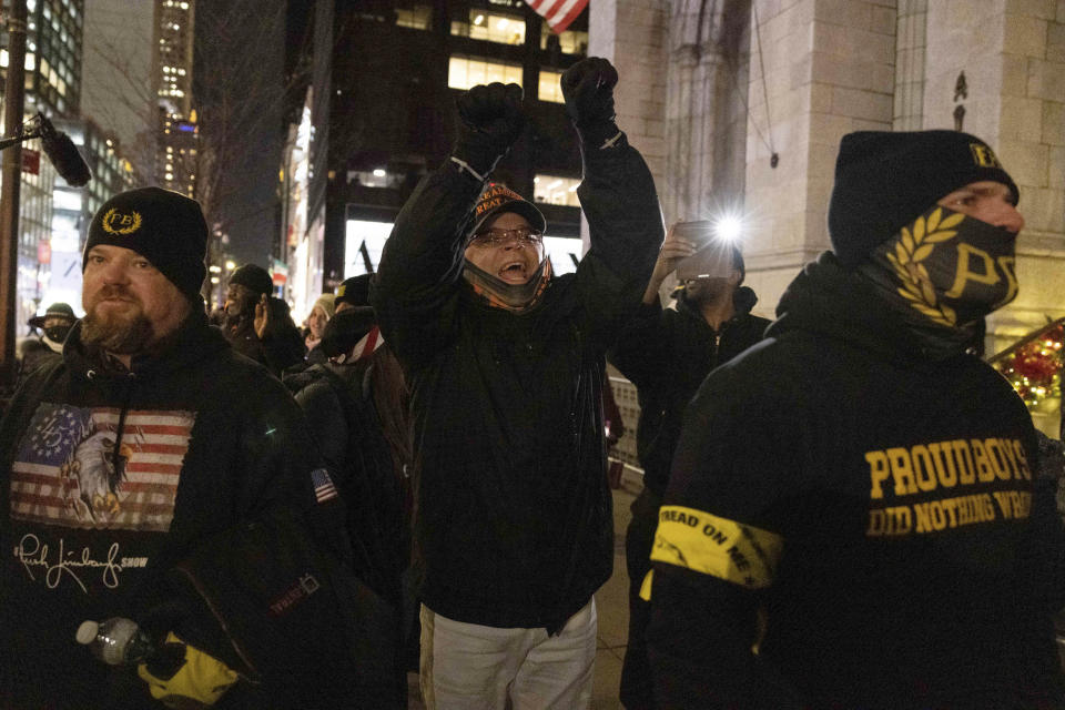 Three men in a crowd of protesters in a street lined with skyscrapers wear Proud Boys yellow insignia, one with his arms raised brandishing an American flag.