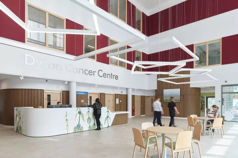 Main entrance atrium for the Dyson Cancer Centre showing the three-storey bright open space. A large painting by Mark Sands is displayed on the far wall and hangs from the ceiling to the first floor. Two people walk through the ground floor space past the reception desk.