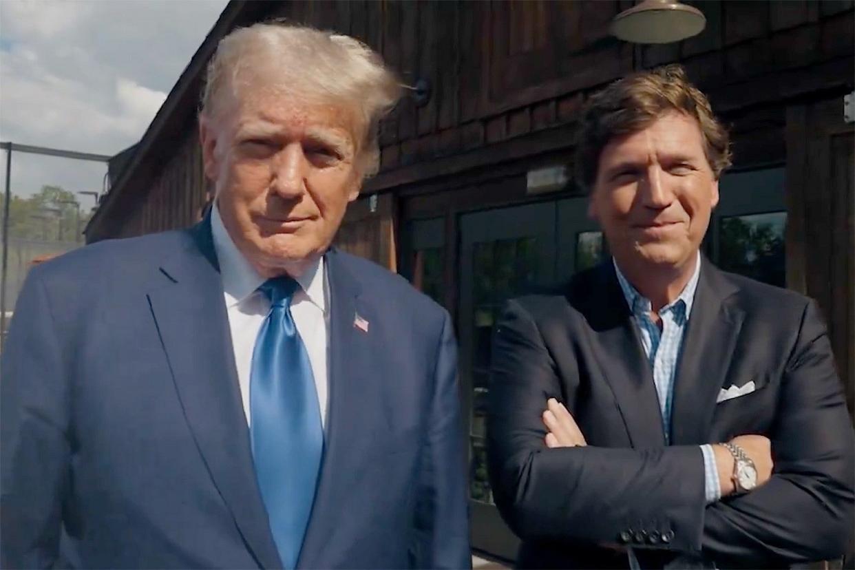 Tucker Carlson with his arms crossed and Donald Trump in a suit stand before what appears to be a barn, looking into the camera. 