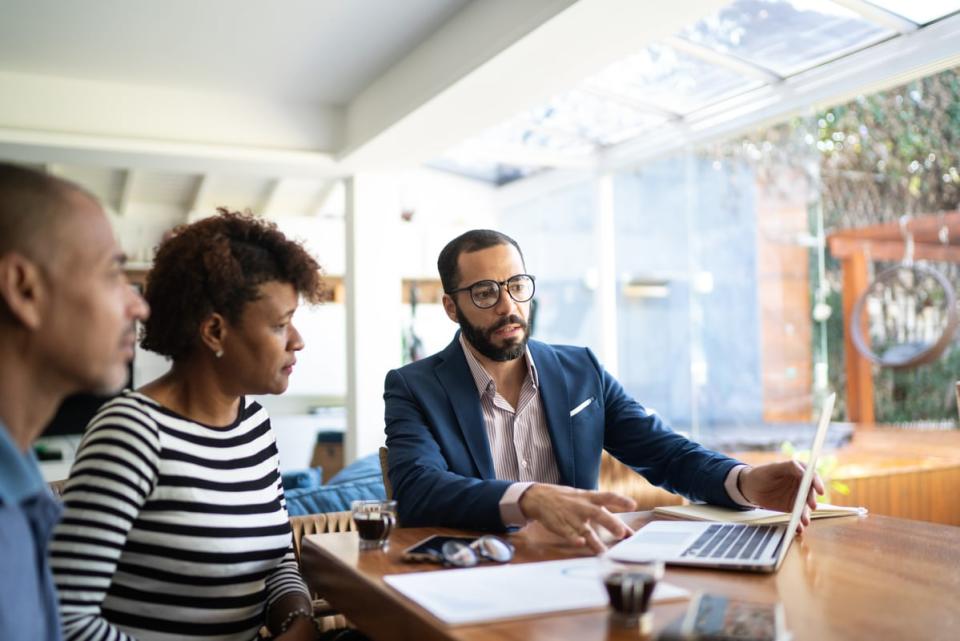 Two people sitting at a table and listening to a person explain something on a laptop computer to them. 