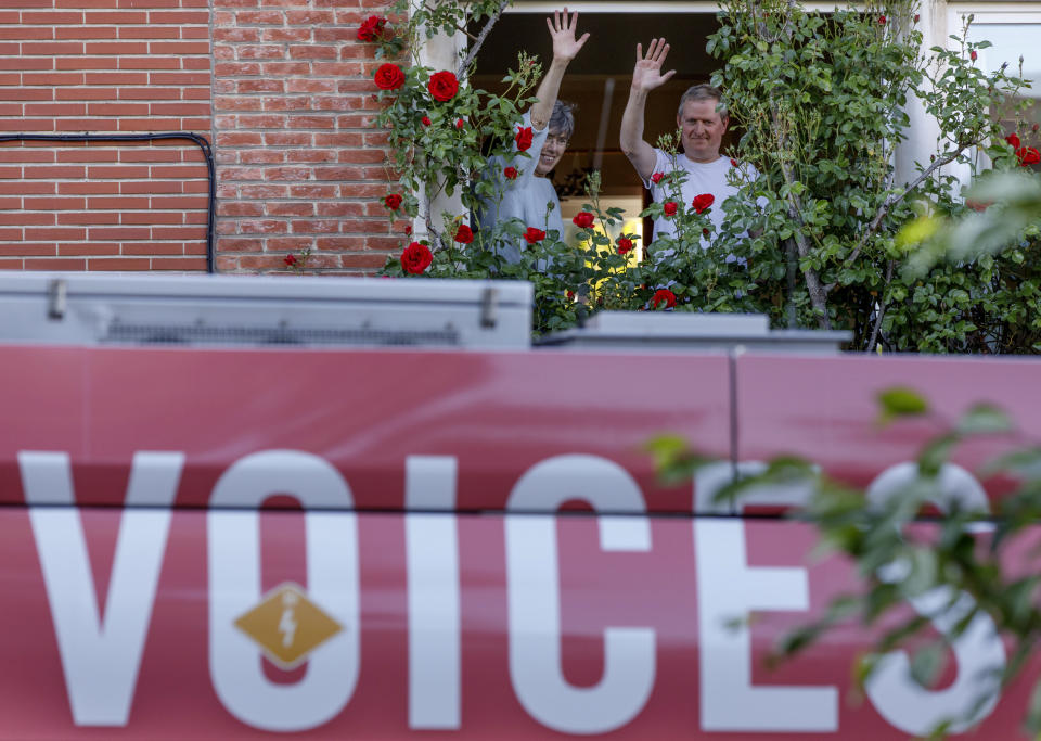 A couple wave from a window as they greet a bus delivering a loudspeaker message from family and friends in Brussels, Wednesday, April 22, 2020. With streets in the Brussels capital mostly devoid of loud traffic and honking horns, a simple piece of emotional poetry can split the air. The public bus company, STIB-MIVB, has been calling on people to send in voice messages, which are now delivered by a special bus driving in a loop to connect all the messages and leave a trail of happiness. (AP Photo/Olivier Matthys)